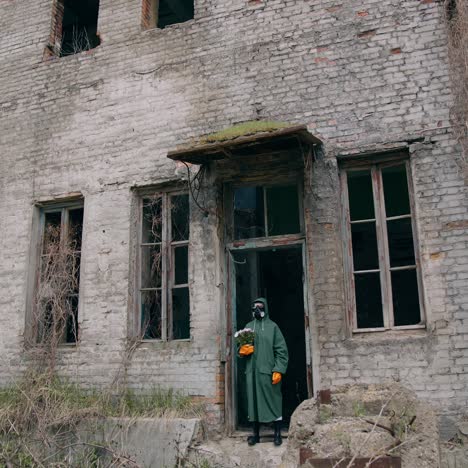 person in safety uniform alone in radiation zone. man in protective suit and gas mask with flowers near abandoned building after the chemical attack