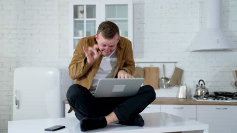 Young man sitting on a kitchen table with a laptop. Nervous freelancer man sitting at table looking at laptop computer screen typing quickly feeling mad.