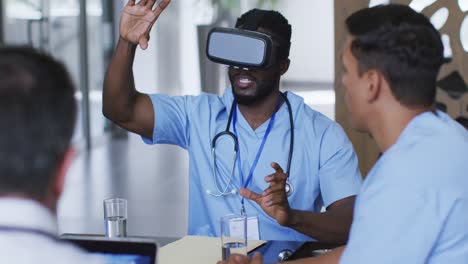 African-american-male-doctor-at-table-using-vr-headset-with-a-diverse-group-of-colleagues-watching