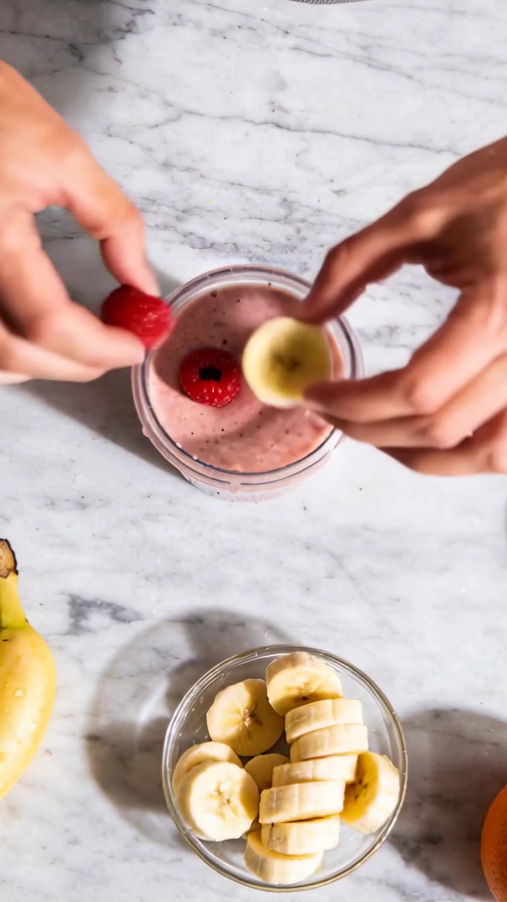 Video verticale: Mani che mettono gli ultimi lamponi mentre finiscono un barattolo di frullato rosa sul bancone, allungandosi per prendere le banane.