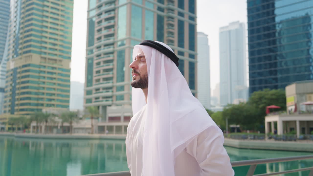 Uomo mediorientale in kandura tradizionale che cammina con sicurezza lungo la baia di Dubai guardando il moderno