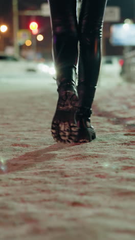 vista en primera persona de una mujer con pantalones de cuero negros y botas negras, caminando por la noche en una calle nevada. luces borrosas de la ciudad y coches son visibles en el fondo