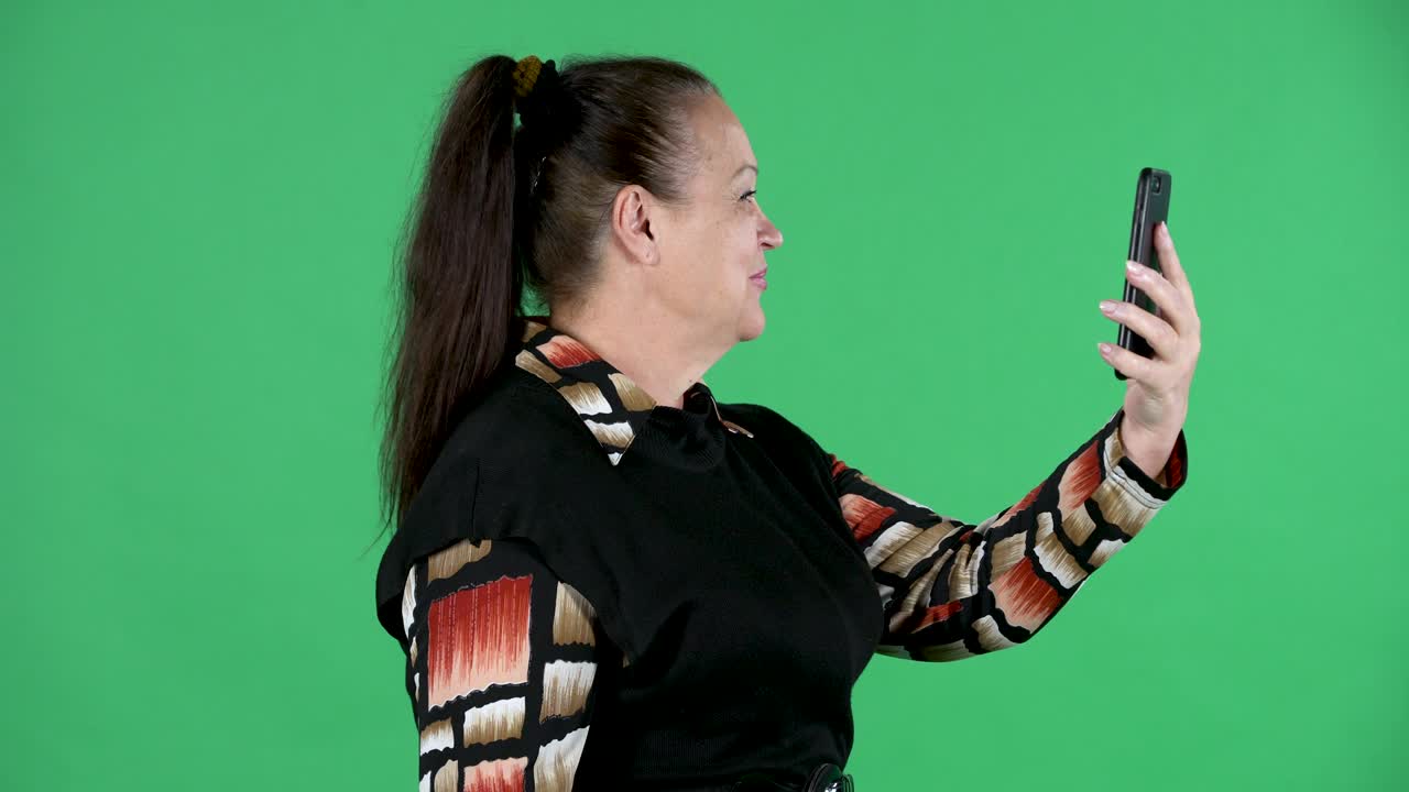 Retrato de mulher idosa conversando em uma videochamada usando seu smartphone. Avó morena com vestido preto em tela verde no estúdio. Vista lateral. Close-up