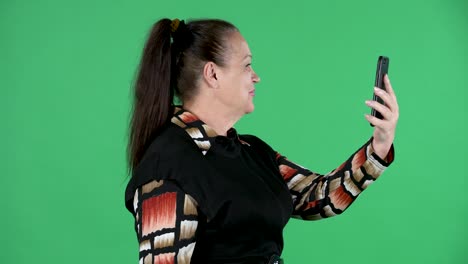 retrato de una mujer anciana hablando en una llamada de video usando su teléfono inteligente. abuela morena en vestido negro en pantalla verde en el estudio. vista lateral. primer plano