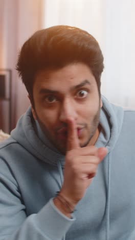 young indian man pressing index finger to lips making silence gesture sitting on sofa couch at home
