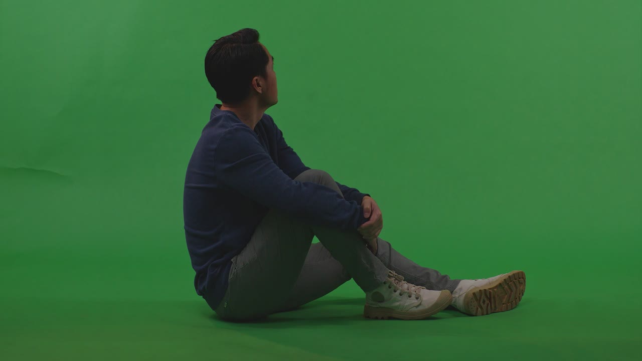 고통스러운 아시아 난민 남성의 몸이 가득 차 있습니다. 주변을 둘러보며 머리를 만지며 전쟁으로 인해 슬프고, 녹색 화면 배경에 바닥에 앉아 있습니다.