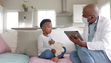 Médico-Afroamericano-Senior-Con-Mascarilla-Usando-Tableta-Hablando-Con-Un-Paciente-Niño,-Cámara-Lenta