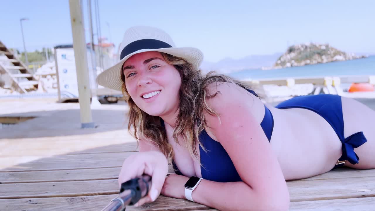 Giovane donna in bikini e cappello sdraiata su un pontile da spiaggia che si fa un selfie