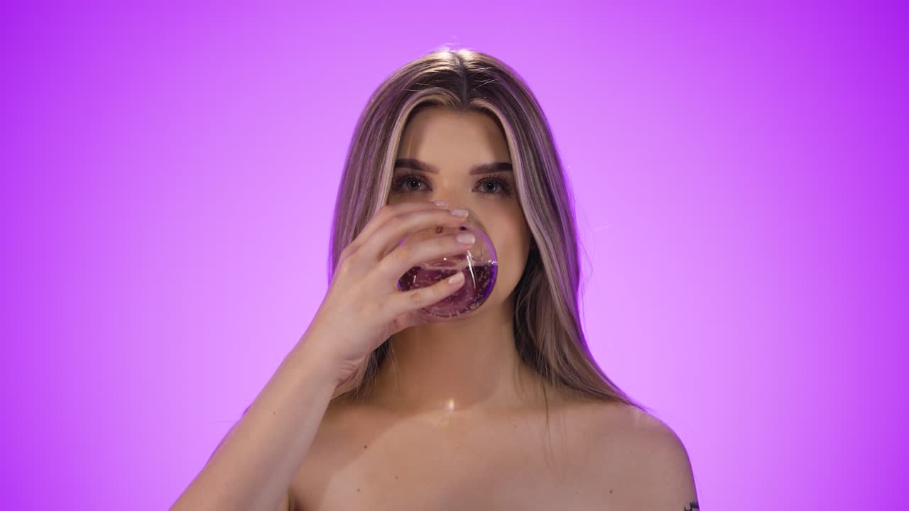 Static shot of a blonde young thirsty woman drinking a glass of water to quench her thirst and looking neutrally into the camera in front of purple background in slow motion