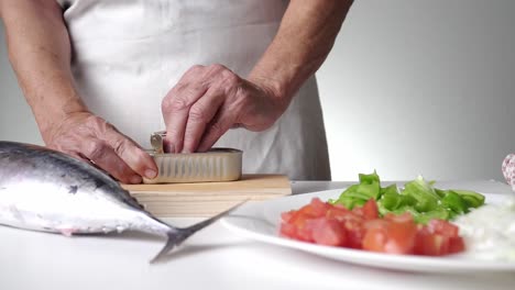 caucasian person with old hands and white apron opening a can of preserves, in the foreground a fish on one side and sliced ​​tomato, green pepper and onion on another side