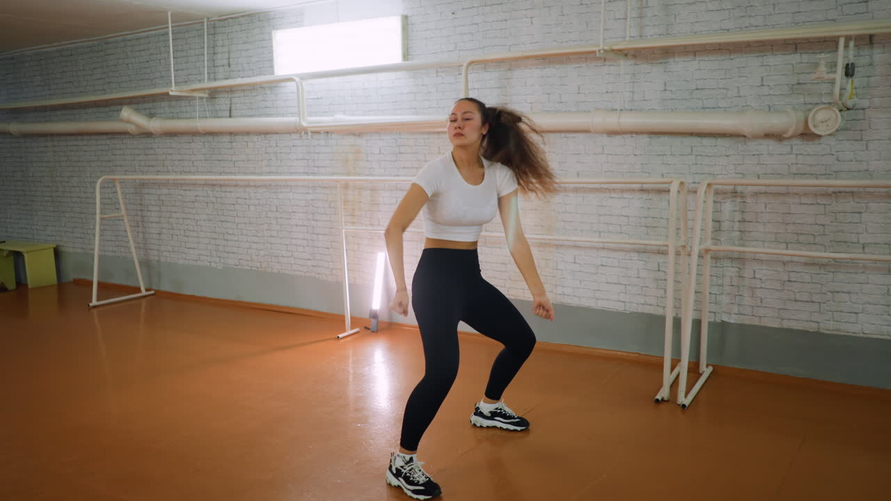 Young girl in casual wear dancing and squatting down near bar and pipe, energetic expressive movement, rhythmic squat and playful choreography in indoor studio with blurred background