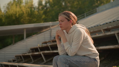 contemplative woman in casual hoodie and jeans sits on rustic stadium bleachers, hands joined together, gazing thoughtfully into distance