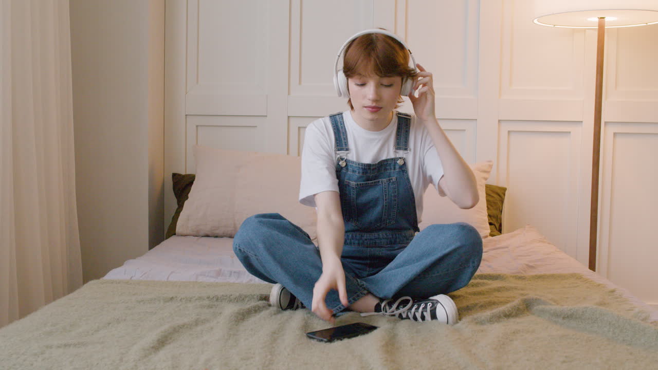 передний вид кавказской девушки, сидящей на кровати, затем она надевает наушники и играет музыку со своего смартфона