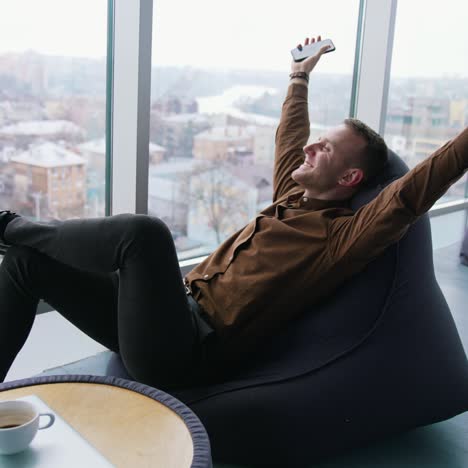 relaxed businessman indoors. happy young man is sitting by the window with stretched arms and having a rest in office on the city view window background