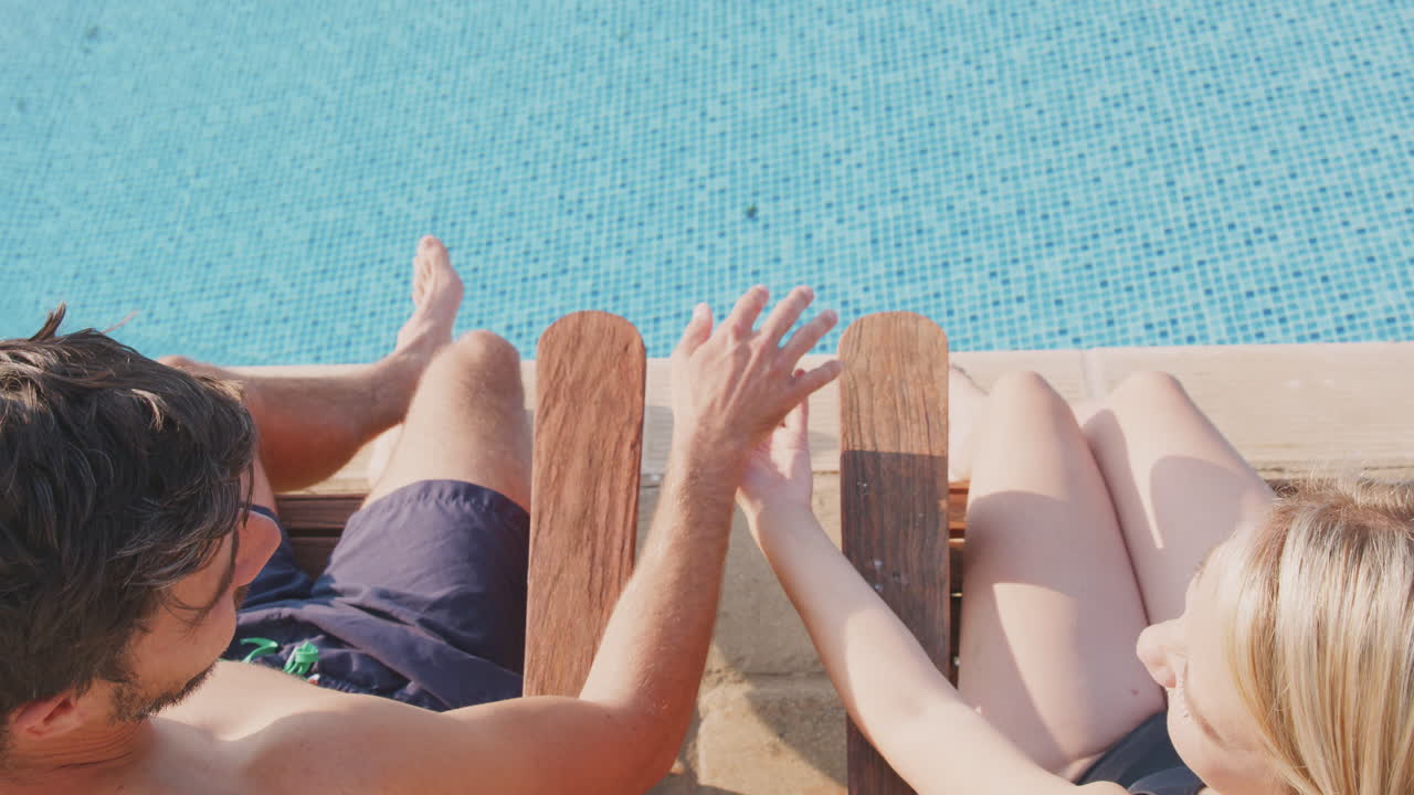 Rear View Of Couple On Summer Vacation Sunbathing On Loungers And Relaxing By Outdoor Swimming Pool