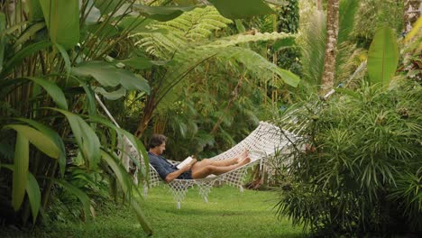 young man lying in hammock reading a book in green jungle paradise surrounded by plants