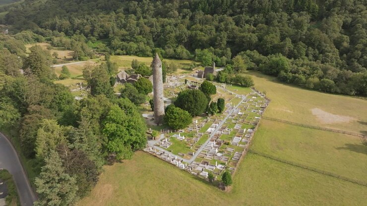 Una veduta aerea mostra una torre circolare in pietra accanto alle rovine di una chiesa e file di tombe, circondata da campi erbosi e una collina boscosa. Glendalough, contea di Wicklow