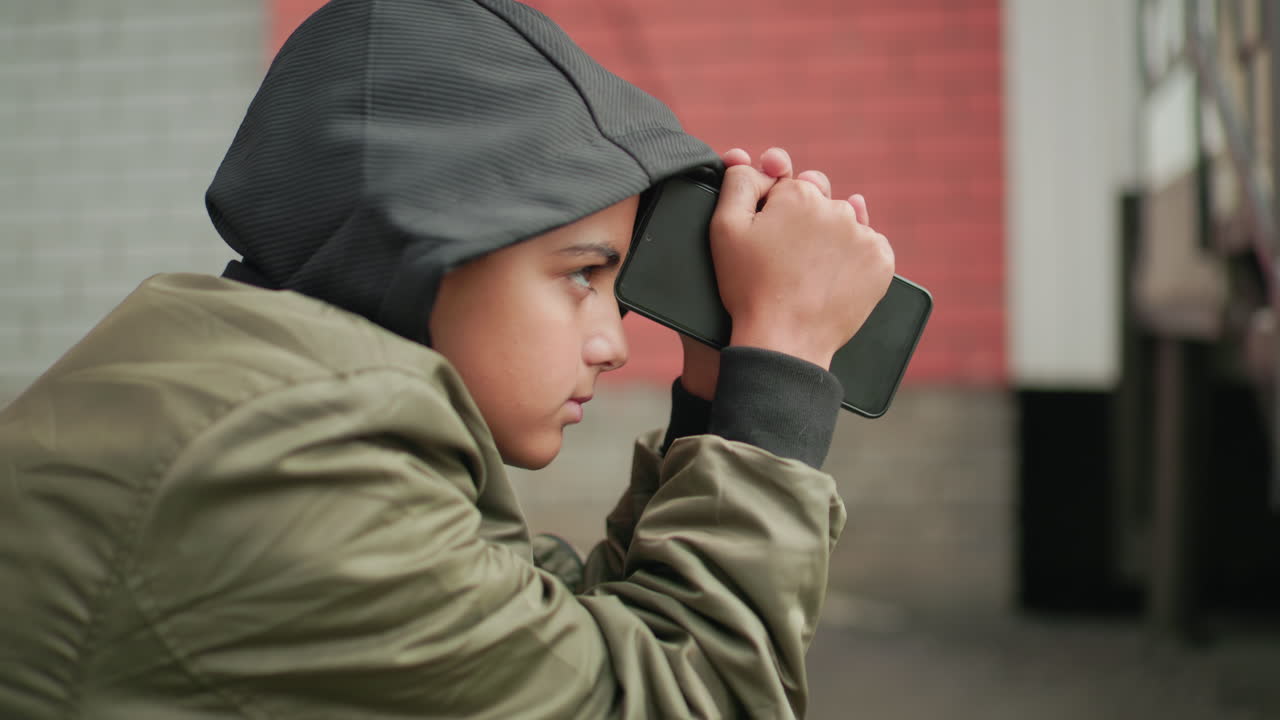 Close up of boy in hooded jacket holding phone against forehead, eyes closed, appearing deep in thought while sitting outdoors near brick wall, conveying emotion and reflection