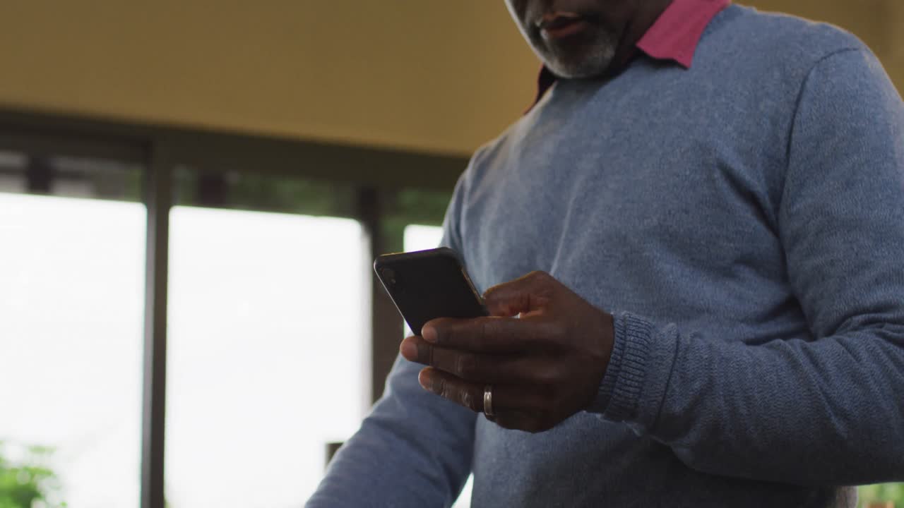 African american senior man standing at counter in kitchen using smartphone