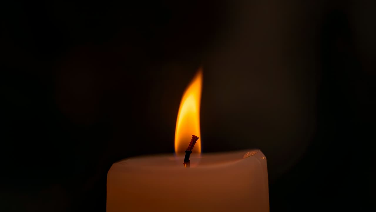 Candela illuminata con stoppino bruciato, fiamma arancione su tavolo in penombra, pozza di cera che riflette il bokeh