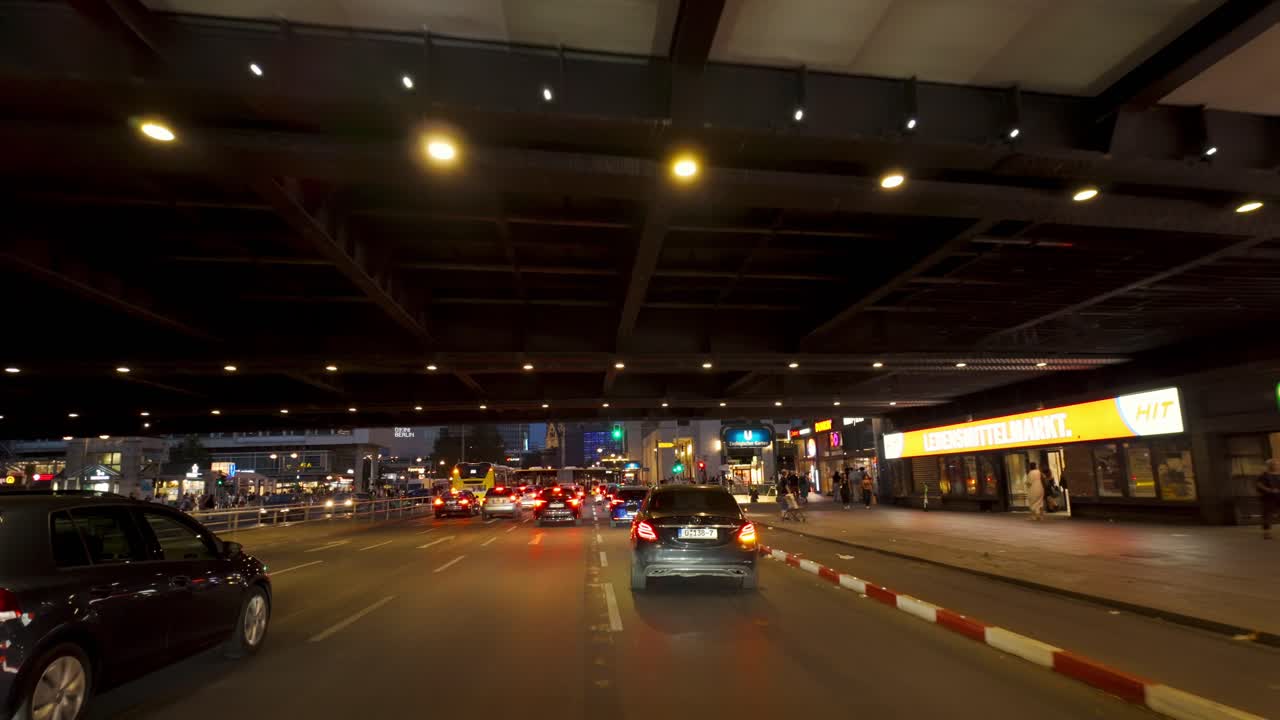 A nighttime driving scene from a car's perspective in Berlin, passing by the Zoologischer Garten area. City lights and modern architecture create a vibrant urban atmosphere.