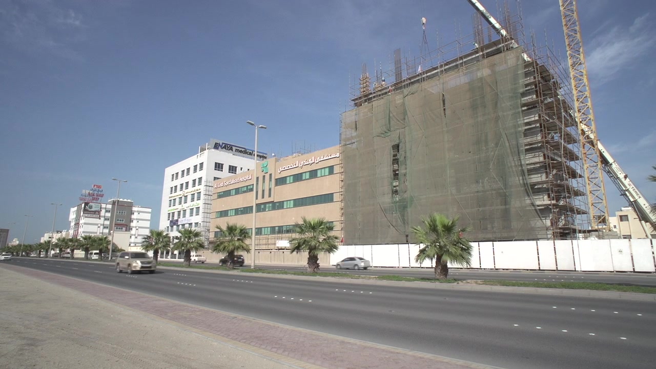 Timelapse callejero con coches cruzando en primer plano del Hospital Especializado Alkindi en Manama, Baréin, con la construcción de la ampliación del hospital y una grúa en la toma.