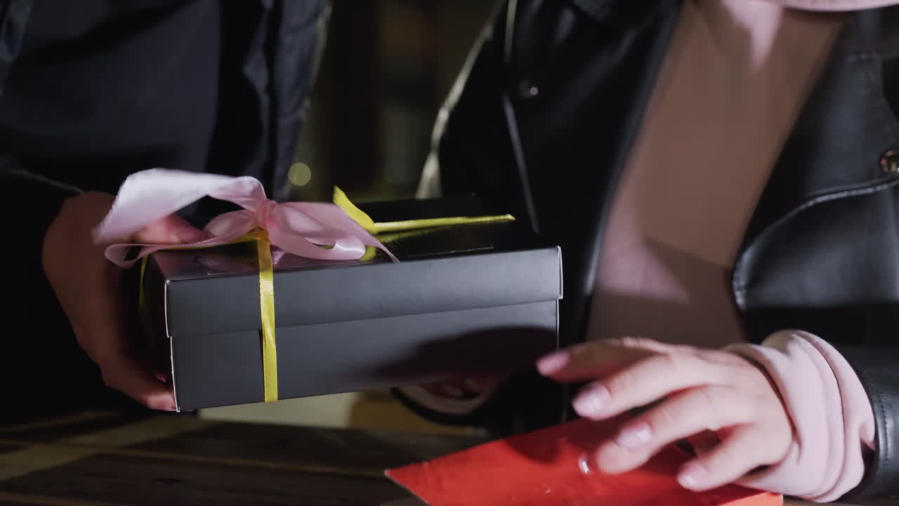 Close up of woman with polished nails reading red cover book at wooden table outdoors as she gently closes it while man holding gift approaches her with soft bokeh light in background at night