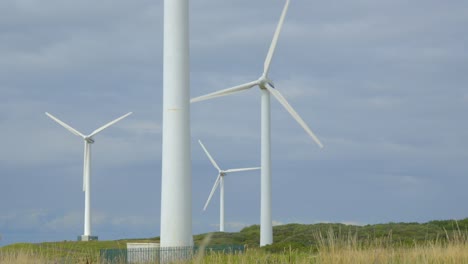 Wind-turbines-through-blurred-grasses-with-rise-up-reveal-clearing-grass-and-showing-windmills-against-cloudy-summer-sky