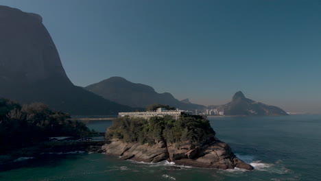 Aerial-approach-of-a-small-island-on-the-coast-of-Rio-de-Janeiro-with-a-construction-on-top-with-the-well-known-city-peaks-of-the-Two-Brothers-and-Corcovado-mountain-behind-on-a-sunny-hazy-afternoon
