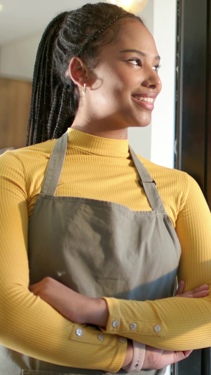 Vídeo vertical: Mulher ouvindo voz na porta, virando e sorrindo na cozinha, com blusa amarela e avental