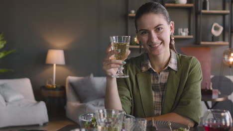 retrato de una mujer bonita sonriente sentada en la mesa de la cena y brindando a la cámara con una copa de vino blanco