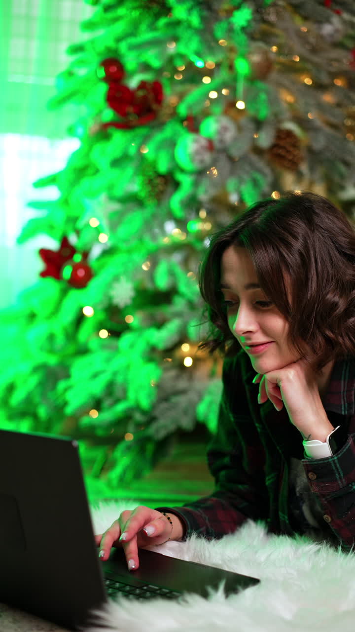 Beautiful Caucasian woman lying at the Christmas tree opens her laptop. Cozy evening at home on holidays. Vertical video.
