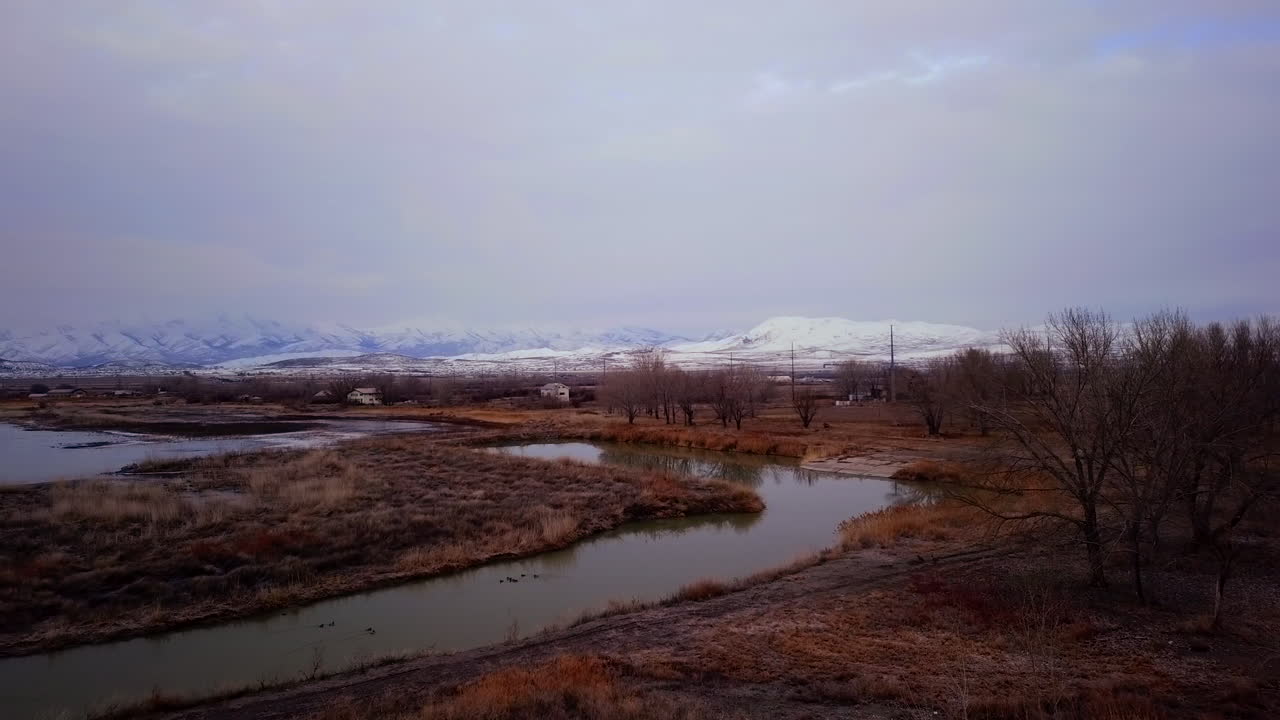 Aerial view of a river flowing into a lake with ducks on the water and mountains in the background at winter