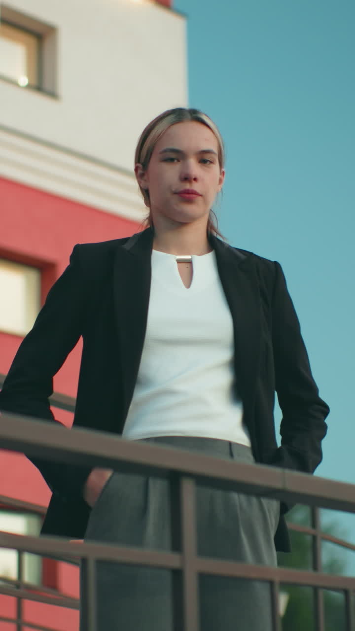 Elegant lady in black blazer and white top poses confidently with hands in pockets outside residence, standing near iron railing with glass windows, red wall, and tree visible in urban background