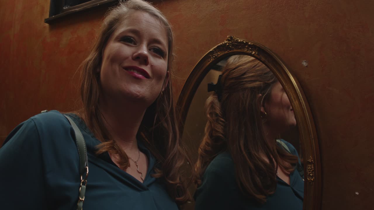 Woman looking in a mirror and putting on lipstick. She looks up to someone off screen and talks while smiling