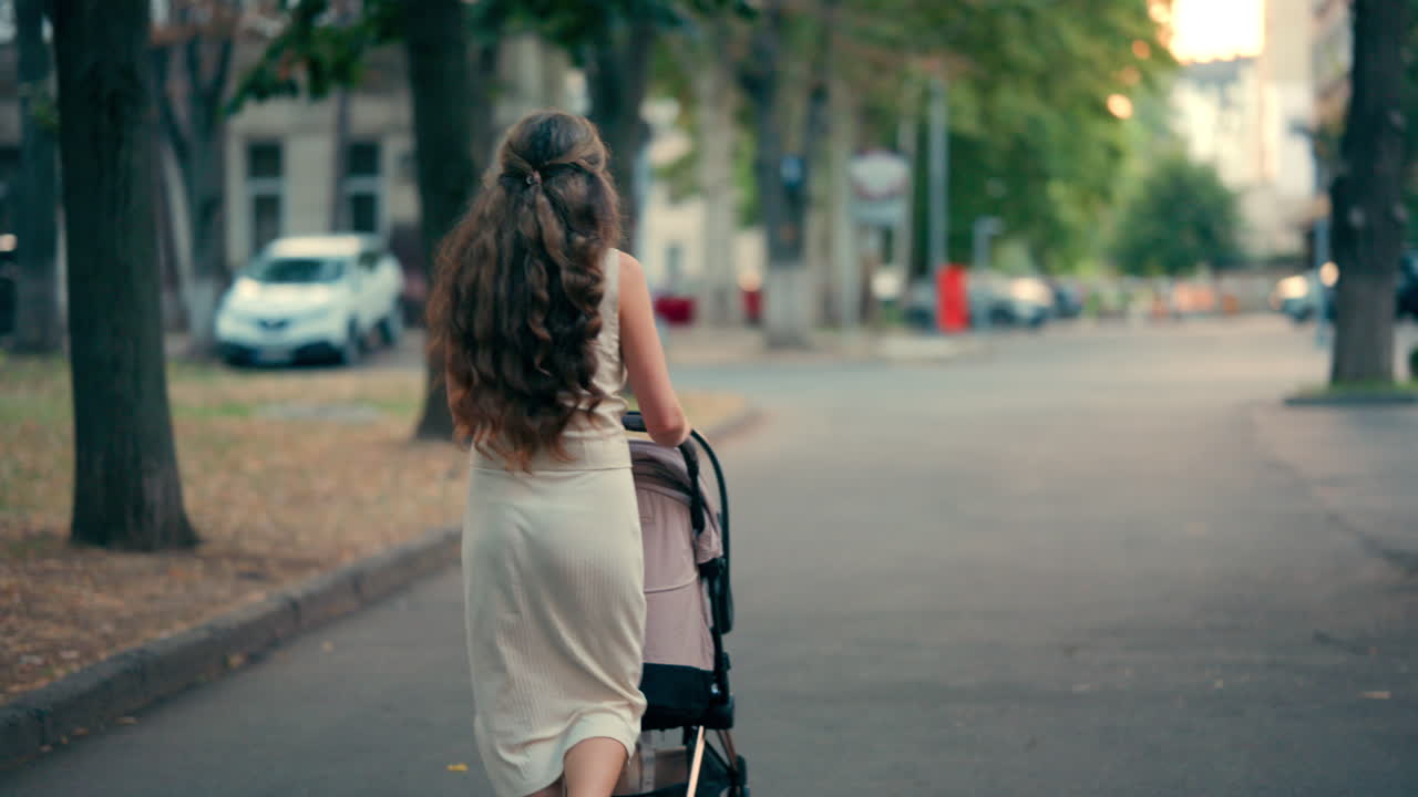 View from the back of a woman with long wavy hair pushing a stroller along a tree-lined park pathway