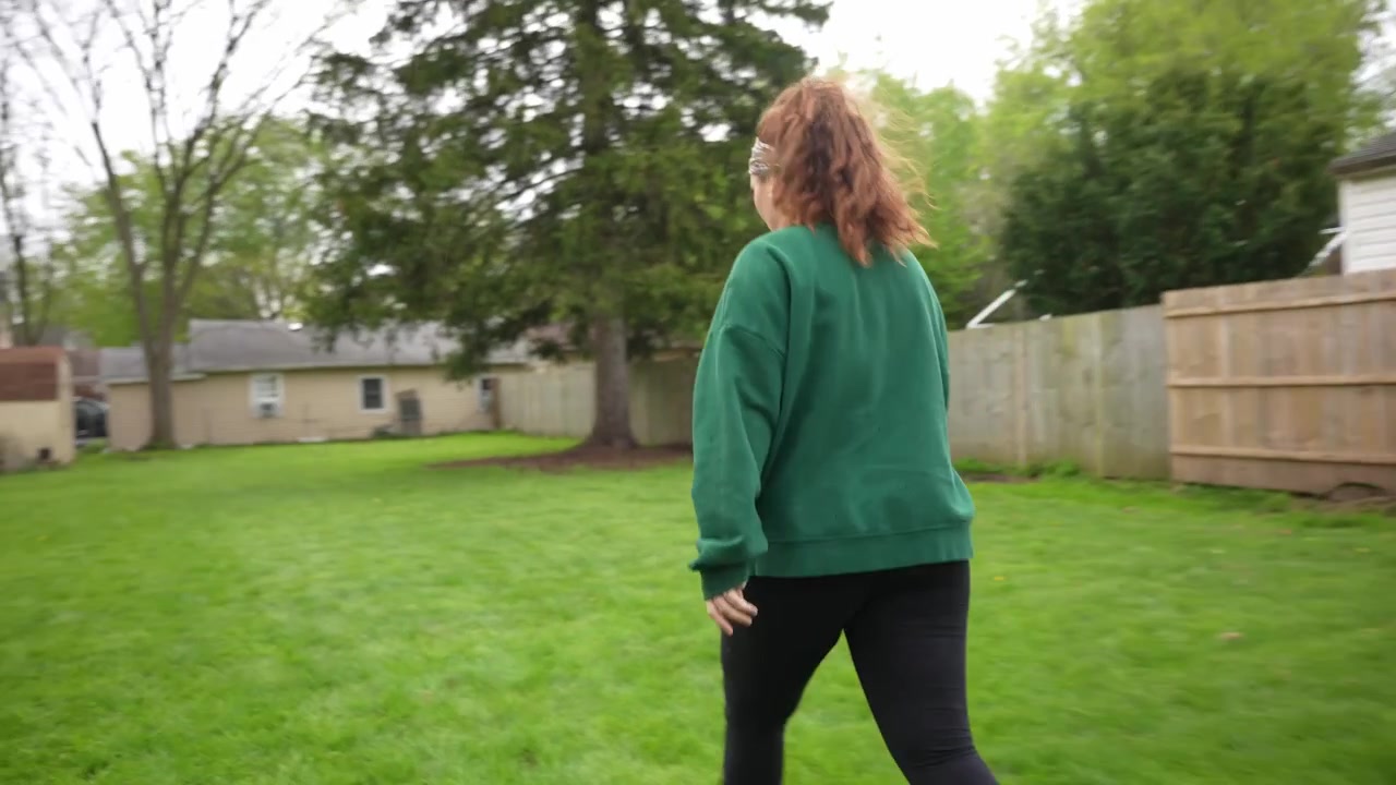 Tomada com câmera na mão de uma mulher correndo por um gramado de quintal, vestindo um suéter verde, com cerca de madeira, árvores e uma atmosfera de casa suburbana casual.