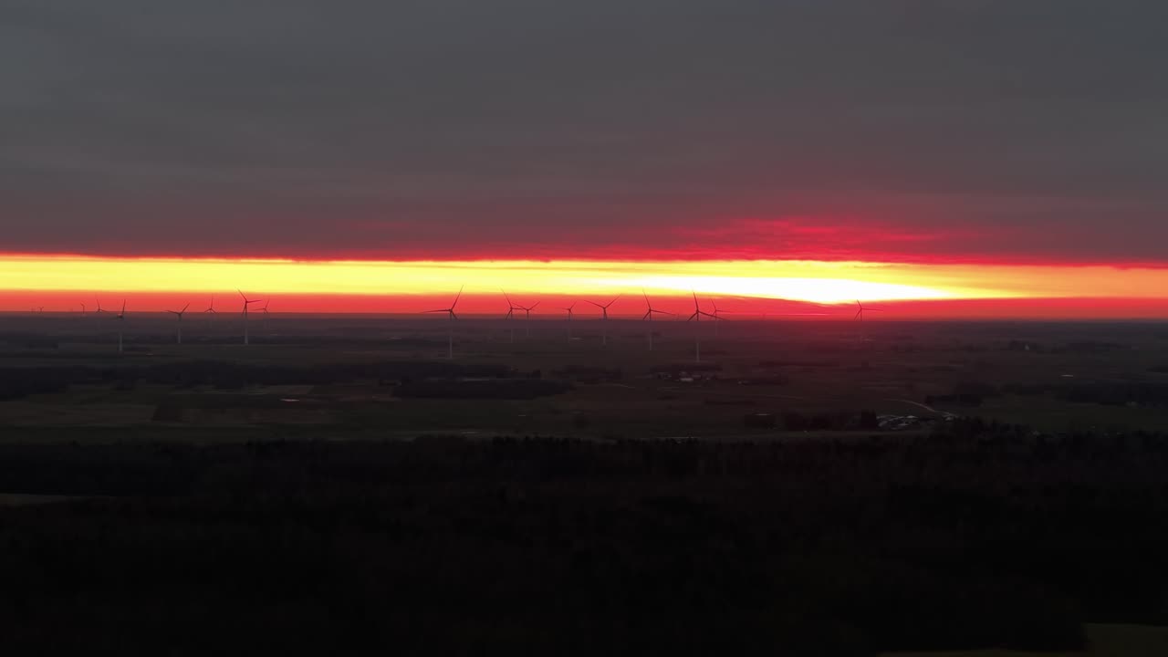 Una vista aerea mozzafiato cattura un vasto parco eolico sotto un cielo drammatico al tramonto. L'orizzonte infuocato illumina il paesaggio, evidenziando le silhouette delle turbine eoliche.