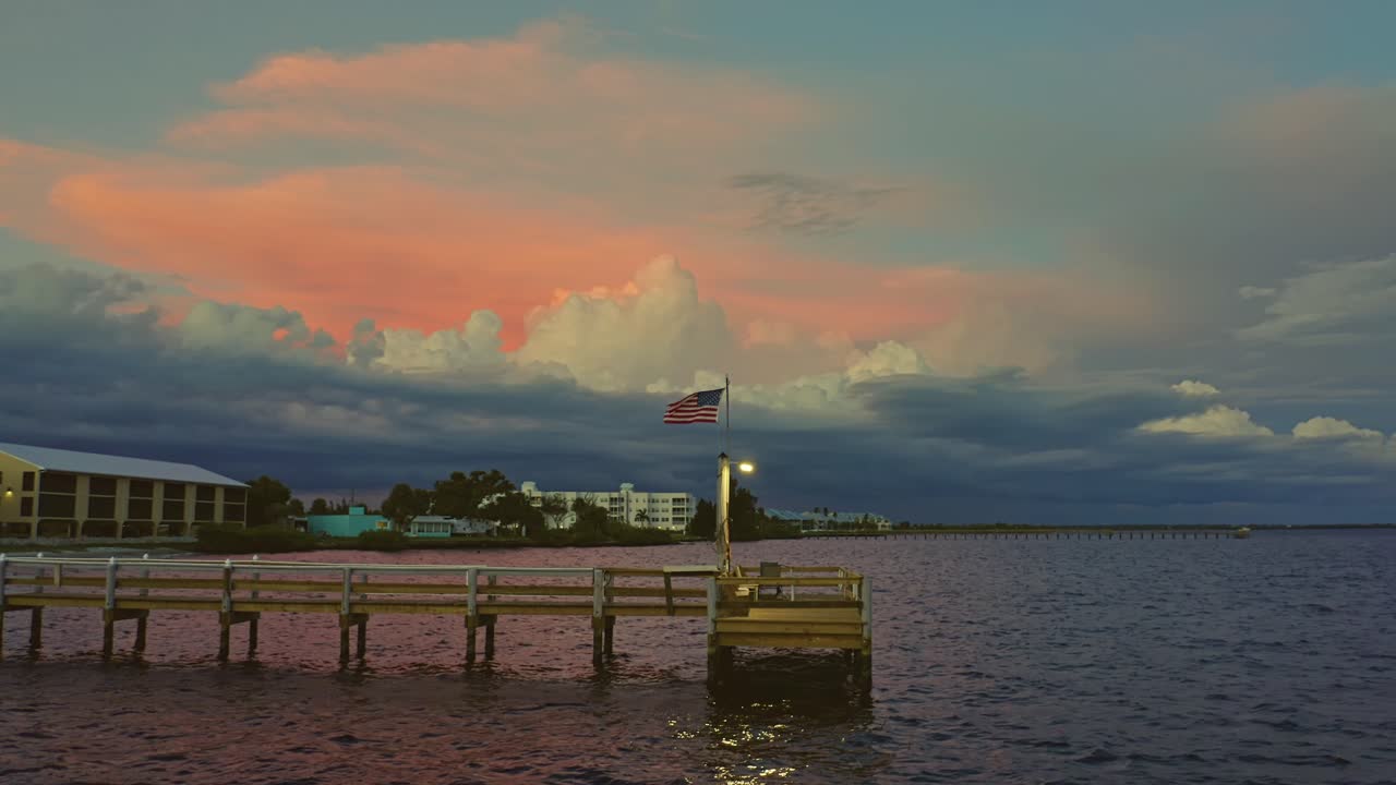 A tranquil pier stretches into the bay as soft sunset hues of orange and pink illuminate the clouds above, while the American flag waves gently in the warm evening breeze