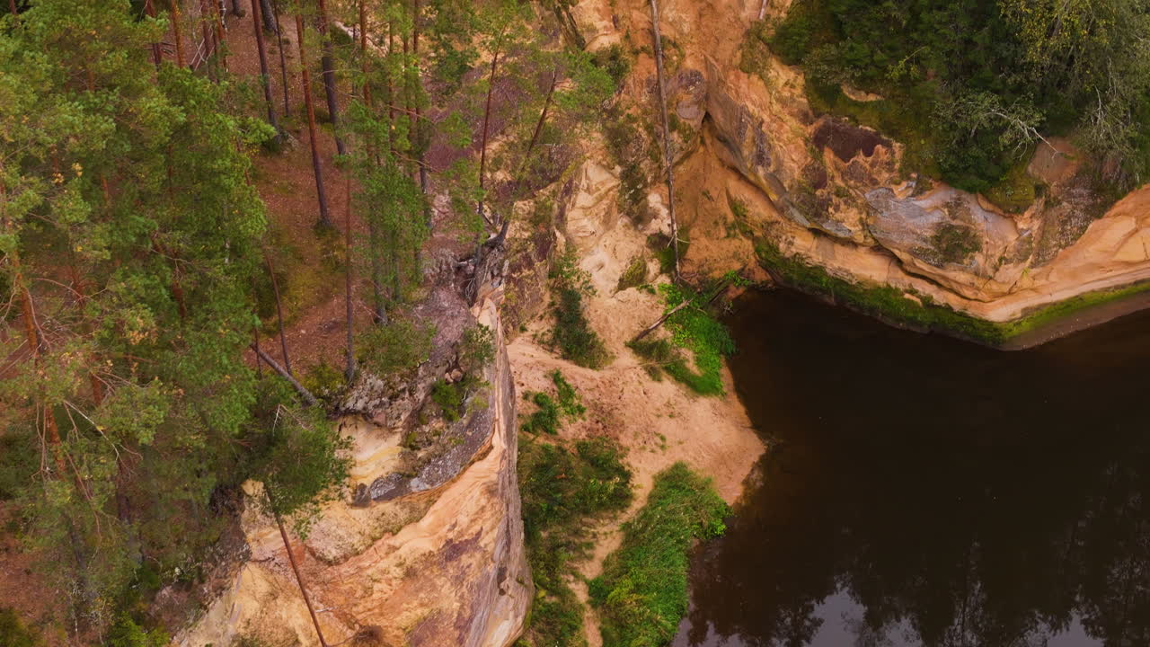 Towering Sandstone Cliffs Rise Above a Dark, Reflective River in Erglu Cliffs, Latvia - High Angle Shot