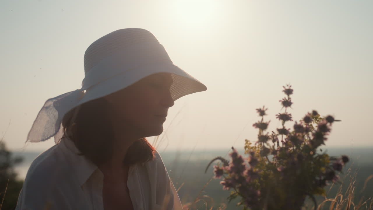 woman in white hat gazing at wildflowers in hand under soft golden sunlight, eyes closed, peaceful expression, sun flare and blurred meadow in background