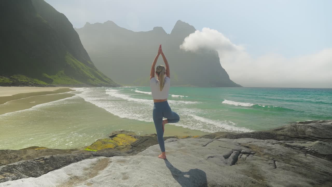 Una donna pratica la posizione dell'albero su una riva rocciosa a Kvalvika Beach nelle Lofoten, Norvegia