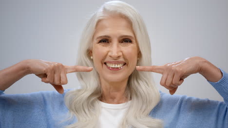 Cheerful-aged-woman-smiling-indoors.-Happy-lady-pointing-to-teeth-in-studio.