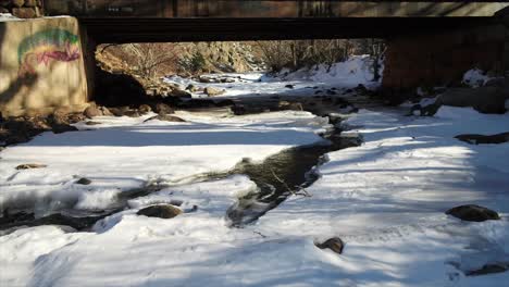 Plätschernder-Bach-In-Der-Schneedecke-Unter-Der-Brücke