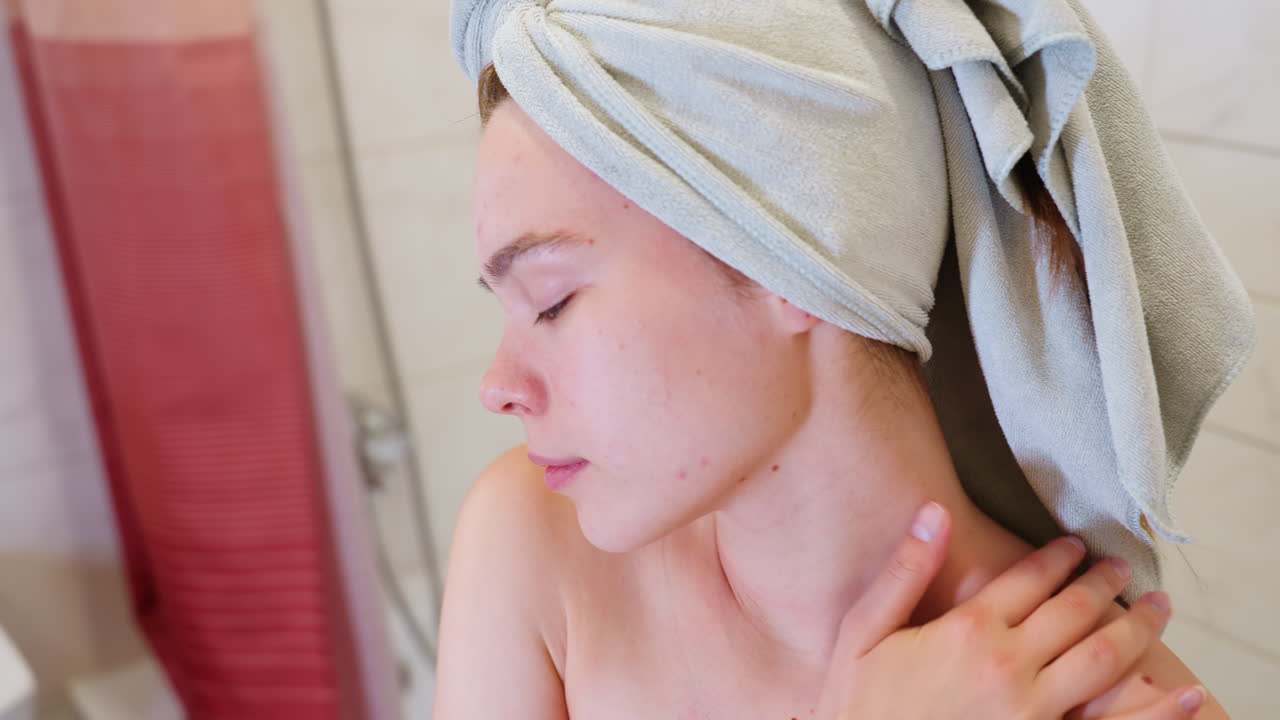 Close-up view of girl massaging neck down to shoulder using hand while wrapped in towel, her hair also wrapped in towel, showing calm facial expression during soothing skincare routine after shower