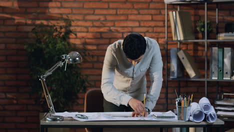 Der-Junge-Architekt-Zeichnet-Einen-Plan-Auf-Das-Große-Blatt-Papier-Auf-Dem-Schreibtisch-In-Seinem-Büro,-Dann-Erhebt-Er-Seinen-Kopf-Und-Lächelt-In-Die-Kamera