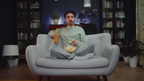 young man watching unexpected movie scene on tv while eating chips and drink soda sitting with crossed legs on couch at home