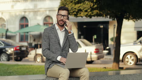 gut aussehender geschäftsmann, der auf seinem handy spricht, während er in der stadt an der wand sitzt und an seinem laptop arbeitet 1