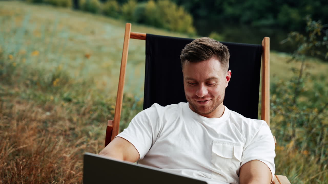 Cheerful Caucasian entrepreneur looks at his laptop. Man working relaxed in the nature backdrop.