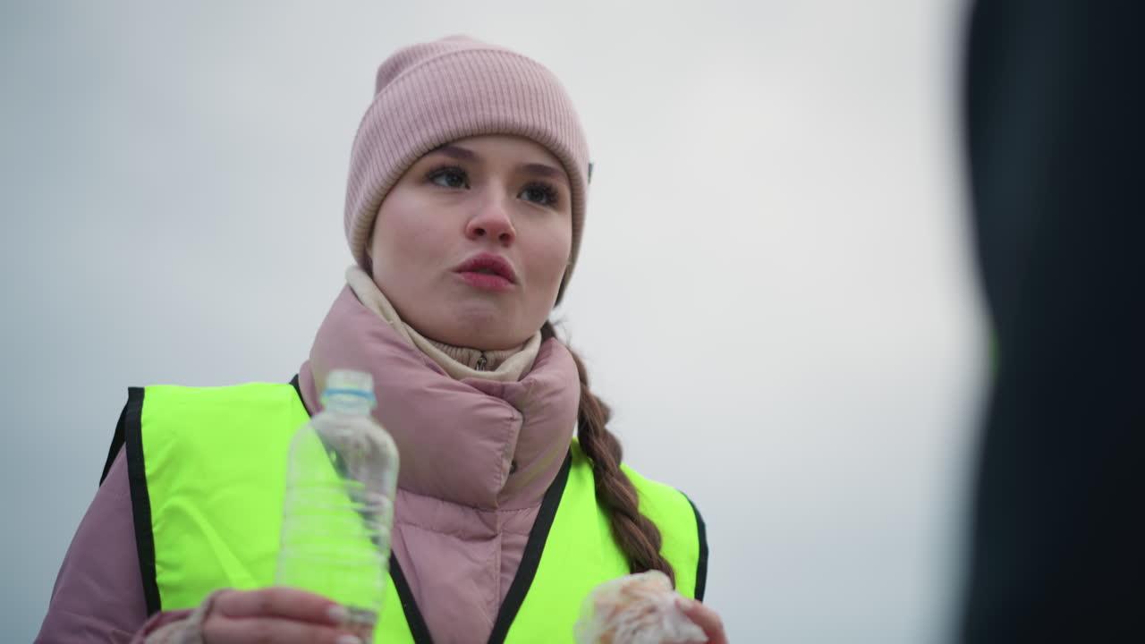 Confident female worker wearing pink beanie, thick scarf, and reflective safety vest holding water bottle and sandwich outdoors on overcast winter day, looking ahead with calm satisfied expression